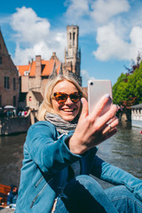 Wall Mural - Beautiful young girl takes selfie photo on the background of the famous tourist destination with a canal in Bruges, Belgium