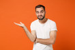 © ViDi Studio - Confused puzzled young man in casual white t-shirt posing isolated on bright orange wall background, studio portrait. People sincere emotions lifestyle concept. Mock up copy space. Spreading hands.