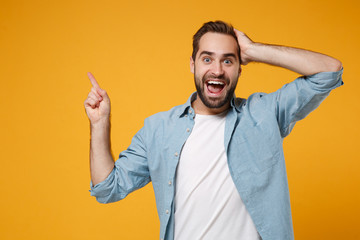 Poster - Excited young man in casual blue shirt posing isolated on yellow orange wall background, studio portrait. People lifestyle concept. Mock up copy space. Pointing index finger up, putting hand on head.