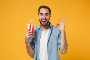 Wall Mural - Excited young man in casual blue shirt posing isolated on yellow orange wall background, studio portrait. People lifestyle concept. Mock up copy space. Holding cup of soda or cola, showing OK gesture.