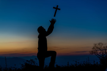 Wall Mural - Silhouette of a young Christian man who is praying on the holy cell in the Bible and raising a cross over a Christian with a light sunset background. Christian concept