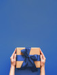 canvas print picture - Female hands hold gift box on deep blue background with copy space for design. Caucasian girl hands holding gift box in craft wrapping paper with fashion color 2020 blue satin ribbon. Vertical.
