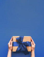 Female Hands Hold Gift Box On Deep Blue Background With Copy Space For Design. Caucasian Girl Hands Holding Gift Box In Craft Wrapping Paper With Fashion Color 2020 Blue Satin Ribbon. Vertical.