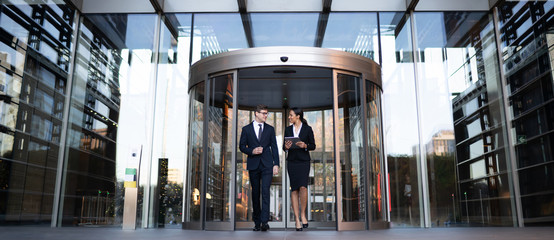 Smiling young coworkers talking on street
Contemporary elegant businesswoman with tablet talking to male colleague while walking out of modern office building with glass revolving doors