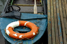 A Lifeboat Moored Near A Wooden Pier. Inside Is A Wooden Oar And A Life Preserver. An Old Motorboat With A Steering Wheel. Poor Working Conditions. Use Of Old Equipment.