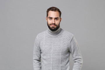 Poster - Handsome young bearded man in gray sweater posing isolated on grey wall background, studio portrait. Healthy fashion lifestyle, people emotions cold season concept. Mock up copy space. Looking camera.