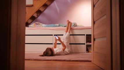 Kid girl of school age, lie on a floor in bedroom texting messages to friends using smartphone having fun before go to bed