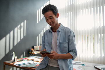 Wall Mural - enjoying his work. portrait of young cheerful asian man using touchpad and smiling while leaning on 