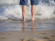 Feet Of A Man Wading In The Waves Of A Beach Reflected In The Sand