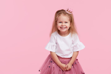 A cheerful little girl with in a tulle skirt and princess crown on her head isolated on a pink background. Celebrating a vibrant carnival for kids, birthday party, having fun