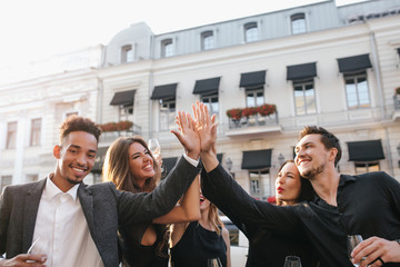 Happy friends clap their hands and laughing, enjoying summer evening in city. Outdoor portrait of inspired african man in trendy jacket drinking champagne with girls n front of old building.