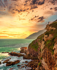 A man stands on a cliff by ocean in sunset