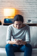 © nenetus - Smiling young woman using her mobile phone while sitting on sofa in the living room at home.