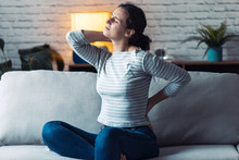 Young Woman With Back And Neck Pain Sitting On The Sofa In The Living Room At Home.