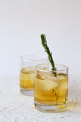 glass of lemonade with pieces of ice and a sprig of rosemary on a white background. two glasses of whiskey and ice on white table.