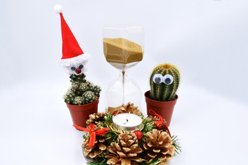 Two green cactus with eyes are standing by the hourglass, golden sand is pouring, time is running a Christmas wreath decorated with cones and a candle.