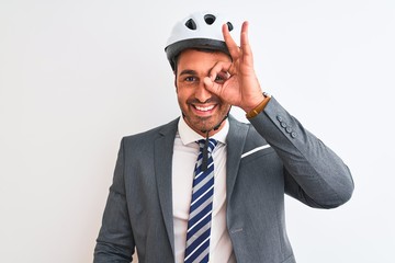 Canvas Print - Young handsome business man wearing suit and tie and bike helmet over isolated background doing ok gesture with hand smiling, eye looking through fingers with happy face.