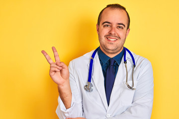 Sticker - Young doctor man wearing coat and stethoscope standing over isolated yellow background smiling with happy face winking at the camera doing victory sign. Number two.