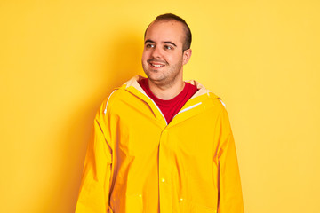 Sticker - Young man wearing rain coat standing over isolated yellow background looking away to side with smile on face, natural expression. Laughing confident.