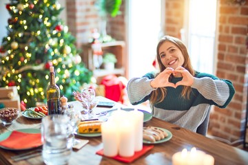 Sticker - Young beautiful woman sitting eating food around christmas tree at home smiling in love showing heart symbol and shape with hands. Romantic concept.