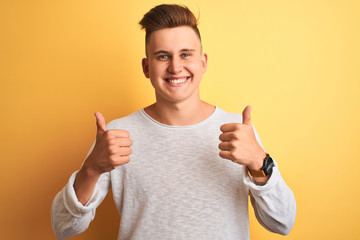 Young handsome man wearing white casual t-shirt standing over isolated yellow background success sign doing positive gesture with hand, thumbs up smiling and happy. Cheerful expression and winner 