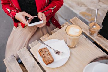 Overhead portrait of girl in red jacket sitting at wooden table with glass of cappuccino. Outdoor photo of young woman holding phone in cafe.