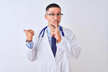 Wall Mural - Young doctor man wearing stethoscope over isolated background asking to be quiet with finger on lips pointing with hand to the side. Silence and secret concept.