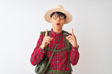 Wall Mural - Chinese hiker man wearing backpack canteen glasses hat over isolated white background amazed and surprised looking up and pointing with fingers and raised arms.