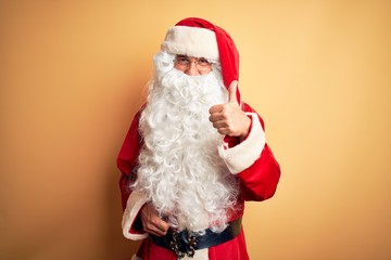 Canvas Print - Middle age handsome man wearing Santa costume standing over isolated yellow background doing happy thumbs up gesture with hand. Approving expression looking at the camera with showing success.