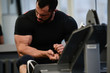 © ruslanshug - sport lifestyle concept of young adult bearded active man sitting in fitness gym looking at his arm muscle during workout break