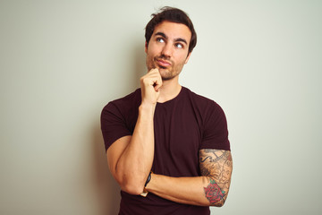 Canvas Print - Young handsome man with tattoo wearing purple casual t-shirt over isolated white background with hand on chin thinking about question, pensive expression. Smiling with thoughtful face. Doubt concept.