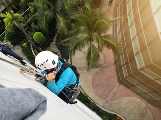 Rope access cleaning worker wearing safety harness hard hat working at height descending on rope performing washes a hospital complex glass wall.