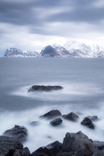 Long Exposure On Rocks And Ocean, With Snowy Landscape In Background. Norway