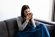 © dianagrytsku - Sick young woman covered with blanket holding cup of tea sitting on sofa couch