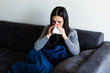© dianagrytsku - Sick young woman sitting on sofa blowing her nose at home in the sitting room