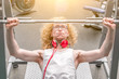 Curly blond man in a white vest lifting barbell with effort.