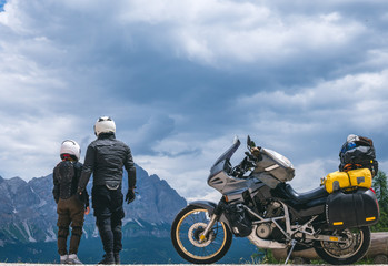 Back view of couple man and girl dressed in helmet and protective equipment for bikers is standing on top of the Dolomites mountains, his touring motorcycle on background, tourism concept, copy space