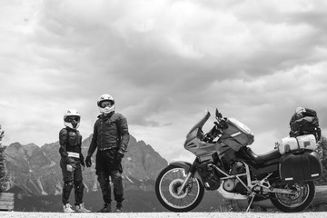 Bikers couple man and girl dressed in helmet and protective equipment for travel, is standing on top of the Dolomites mountains, his touring motorcycle on background, black and white, copy space.