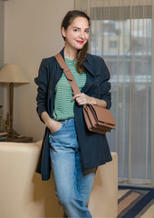 Portrait of mature beautiful fashionable woman wearing a shoulder bag and a trench coat, looking camera over home background, indoors.