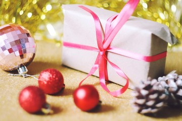 Christmas home decoration. Wrapped gift box, pink, red balls, pine cones on a shiny golden background. New Year present with bow ribbon. Winter holidays atmosphere. Selective focus photo