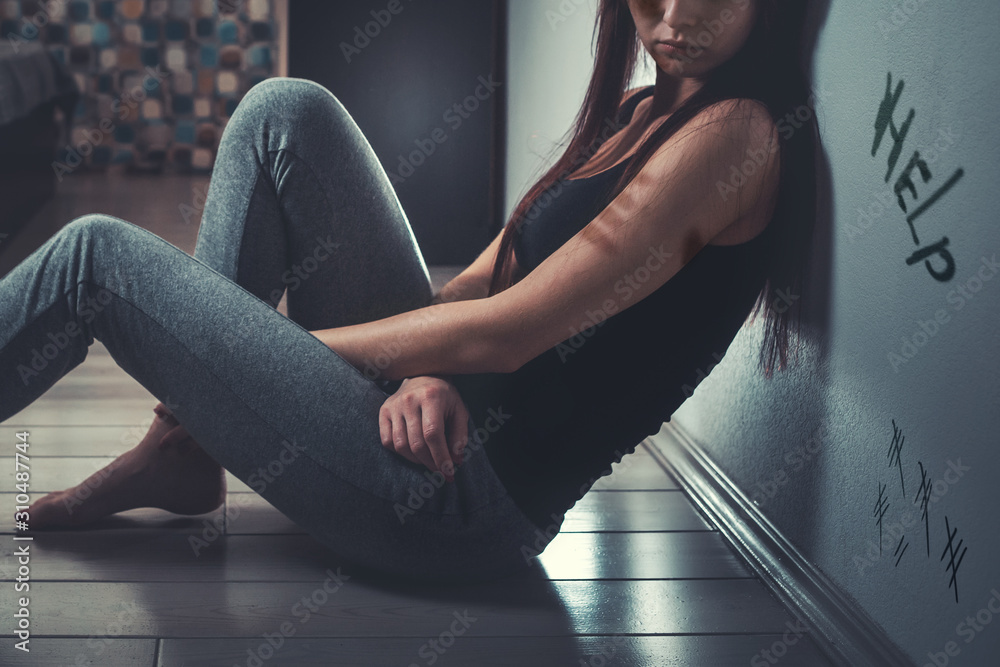 The concept of domestic violence. A sad young woman with a bruise on her arm and under her eye, sitting on the floor in a dark room. In the background, a curtained window. The inscription Help - obrazy, fototapety, plakaty 