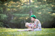 Young mother with green short hair is sitting on green grass, smiling, looking at her toddler son on her laps. Mother's day concept