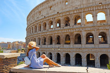 Travel Woman In Romantic Dress And Hat Sitting And Looking On Coliseum, Rome, Italy. Beautiful Tourist Girl With Backpack Near Colosseum. Young Woman Enjoy Summer Italian Vacation In Europe.