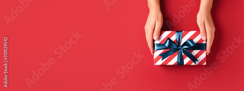 Female's hands holding striped gift box with blue ribbon on red background. C...
