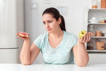 Wall Mural - Making Right Choice. Chubby girl sitting at kitchen choosing between doughnut and apple concerned