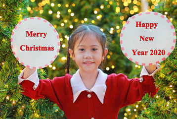 Wall Mural - Portrait of little girl in red dress holding circle label with text Merry Christmas and Happy New Year 2020 in winter season festival against pine tree background.