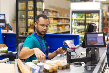 Wall Mural - Focused African American cashier scanning goods at checkout. Concentrated young man in eyeglasses at workplace. Shopping concept