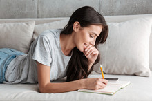 Image of young woman writing down notes in diary book on sofa at home