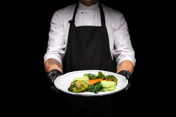 male cook in a black apron holds a plate with cooked bird fillet