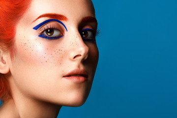 Beautiful girl with colored in red hair and blue eyeliner makeup isolated on blue background